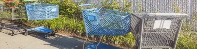 Two grey and two blue abandoned shopping carts sitting on sidewalk with weeds and building wall in the background