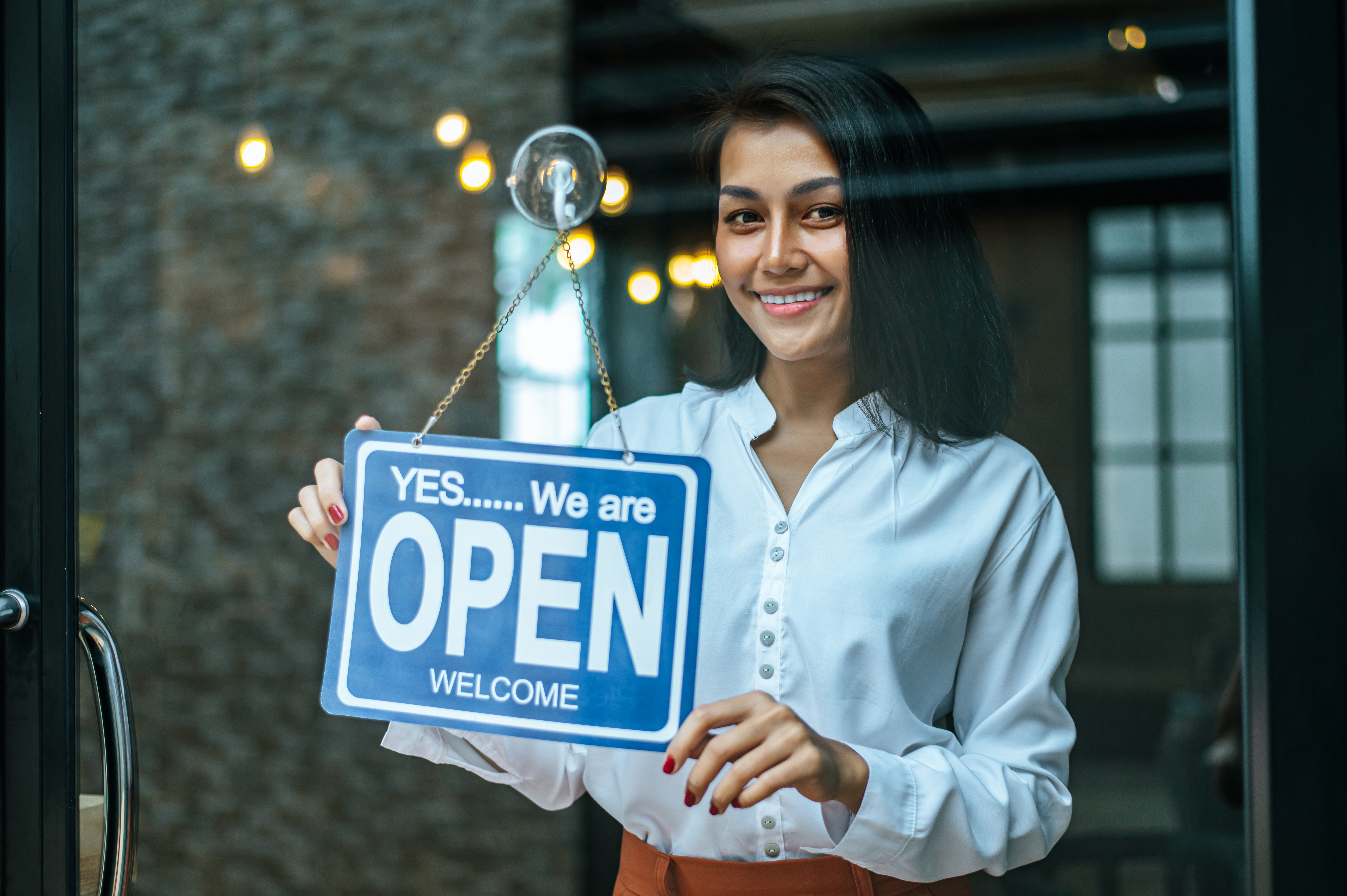 Woman with brown hair  wearing blue shirt smiling  and holding an open sign behind a glass storefront window